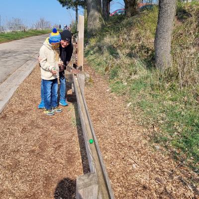 Ein Junge und eine Frau stehen draußen vor einer großen Murmelbahn auf Holz. Sie beobachten wie eine Kugel rollt 