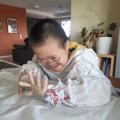 Ein Junge im Malerkittel schaut grinsend seine Hände an. Diese sind in durchsichtigen Handschuhen. 