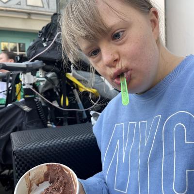 Eine junge Frau schaut in die Kamera, sie hat einen Eisbecher in der Hand und einen Eislöffel im Mund