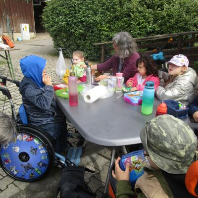 6 Kinder mit und ohne Rollstühle beim Vespern mit ihrer Lehrerin um einen Tisch im Freien auf dem Gelände der Karlshöhe Ludwigsburg. Im Hintergrund Büsche und Stall.