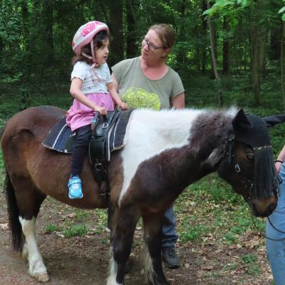 Ein kleinwüchsiges 8jähriges Mädchen mit Helm auf einem dunkelbraun-weiß gefleckten Pony, das nach rechts schaut. Hinter dem Pony, dem Kind zugewandt, eine Frau.