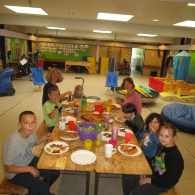 2 Lehrerinnen und 6 Kinder im Grundschulalter um 2 Biertische in der E-Bau-Aula der AHWS. Auf dem Tisch Teller mit Gegrilltem und Salate. 