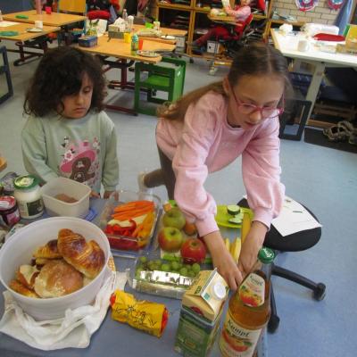 Zwei Mädchen hinter einem reichhaltig gedeckten Tisch mit Obst, Gemüse, frischem Gebäck, Saft und Joghurt. im Hintergrund Schülertische und ein Mädchen im Therapiestuhl.