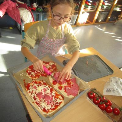 Ein Mädchen verteilt im Klassenzimmer Tomatenstücke und Salami auf seinem Pizzateig.