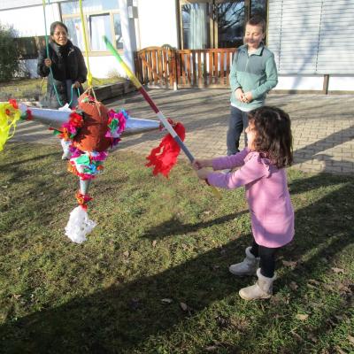 Ein Mädchen schlägt im Freien vor der Schule mit einem Stab auf eine mexikanische Pinata, eine Frau und ein Junge schauen zu