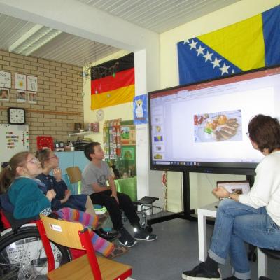 Kinder im Halbkreis vor einem Legamaster, auf dem die Lehrerin gerade Cevapcici zeigt. Oberhalb im Klassenzimmer deutsche und bosnische Flagge.