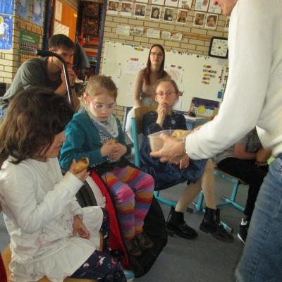 3 Kinder im Stuhlkreis nehmen vom bosnischen Gebäck, das ihre Lehrerin anbietet. Im Hintergrund ein Student und eine Erzieherin.